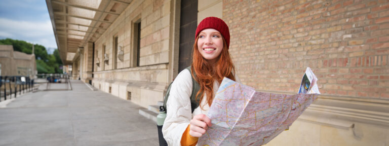 Adventurous redhead girl walks in town with paper map, explores city as tourist, looks for popular tourism attractions, looks around excited and smiles